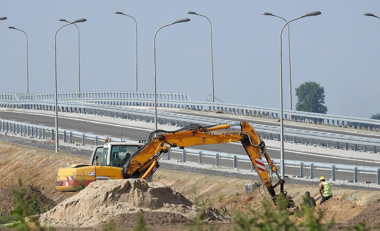 מחפר עובד בסוללת עפר לצד כביש חדש, עם מעקות ועמודי תאורה